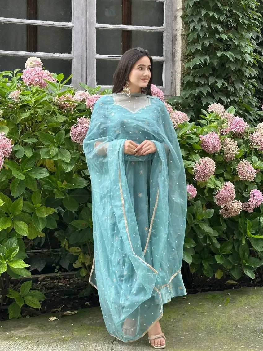 Woman in a light blue saree standing in front of pink flowers and greenery