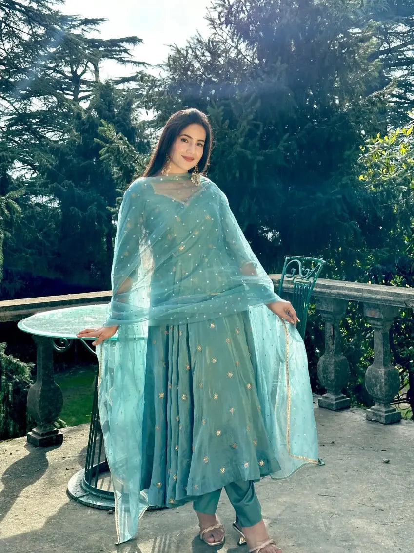 Woman in a light blue traditional outfit standing outdoors with greenery in the background