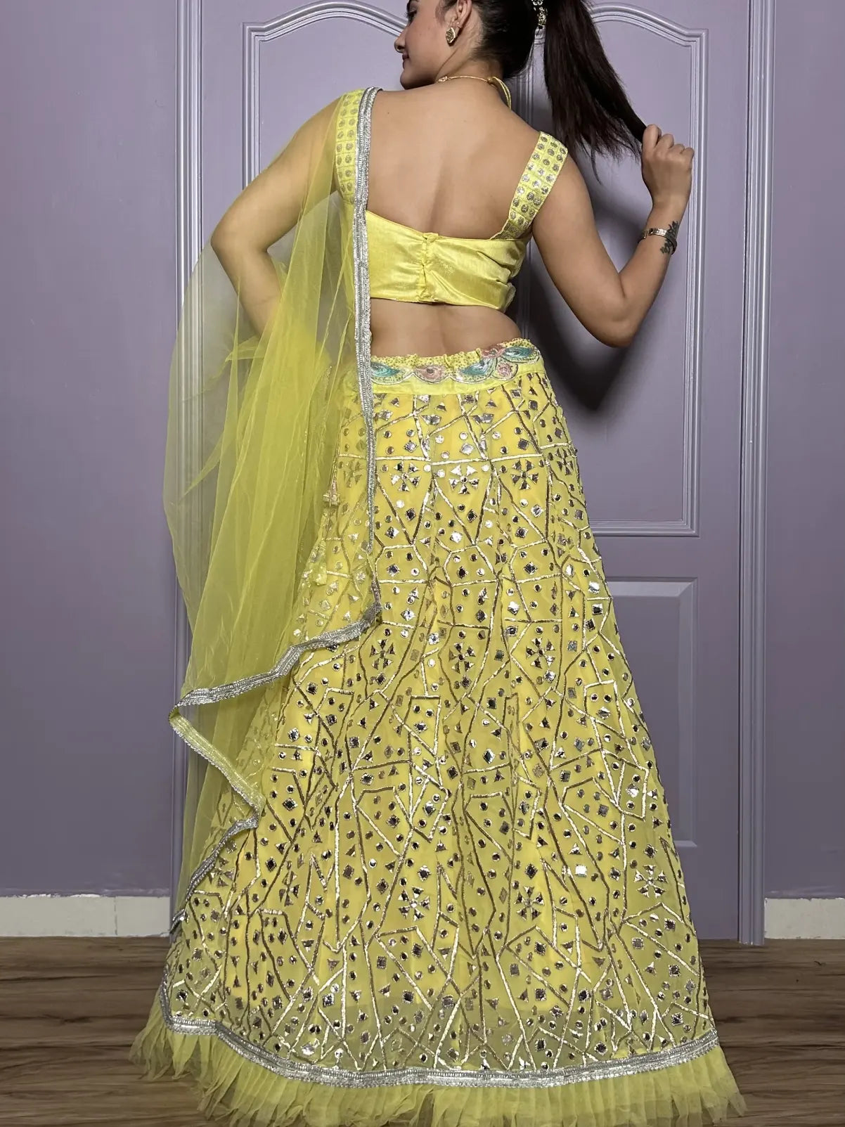 Woman wearing a yellow embroidered lehenga with a matching top against a purple wall.