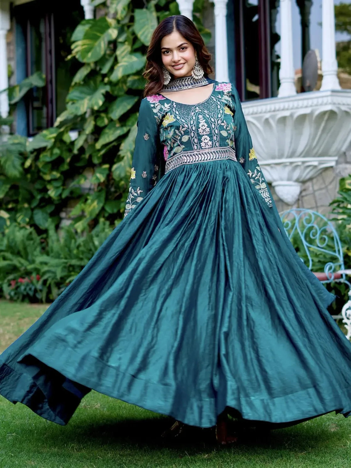 Woman in a teal embroidered dress standing outdoors with greenery and a decorative structure in the background.