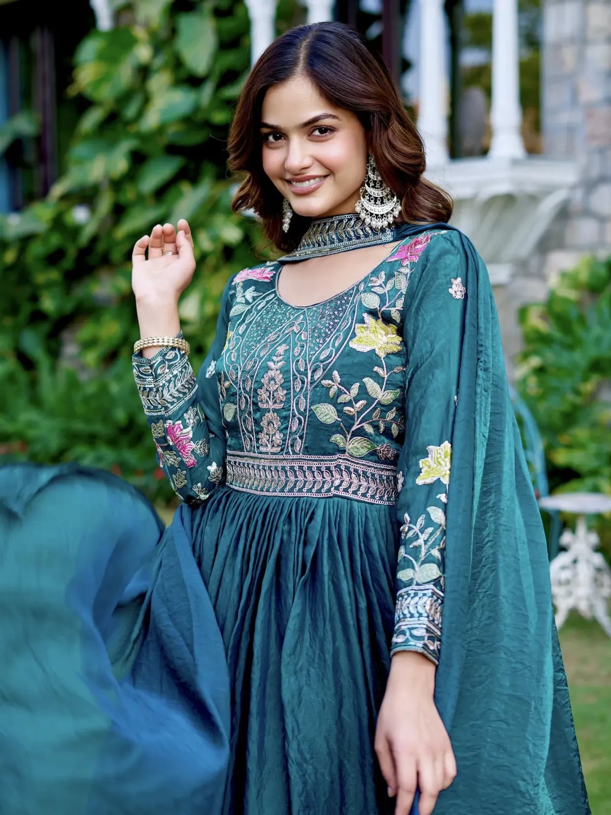 Woman in a teal embroidered traditional outfit with a floral design, standing outdoors.