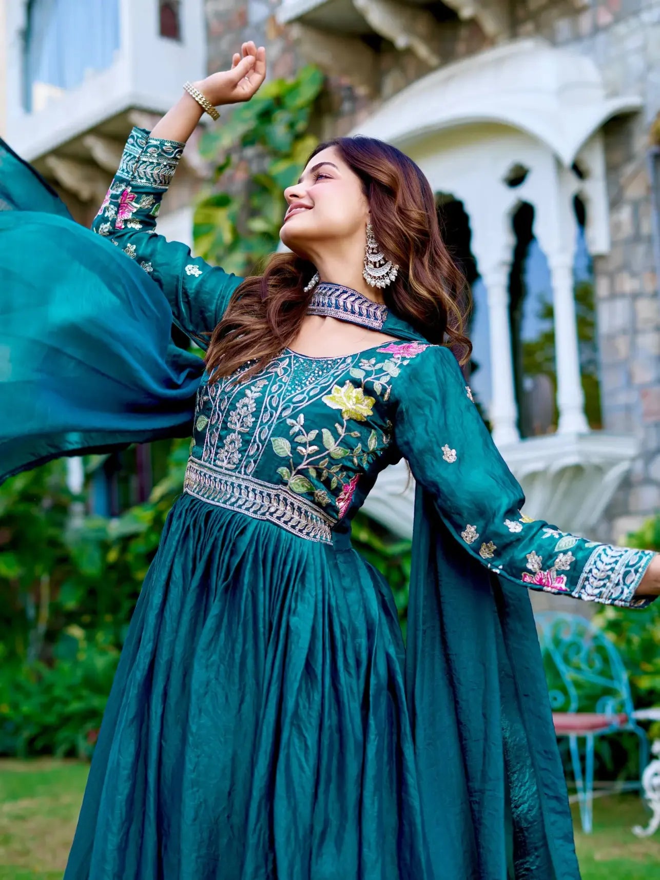 Woman in a teal embroidered outfit standing outdoors with a decorative building in the background