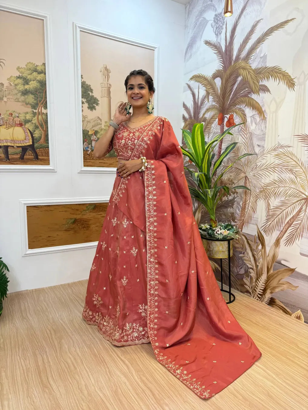 Woman in a red saree standing indoors with decorative wall art and plants.