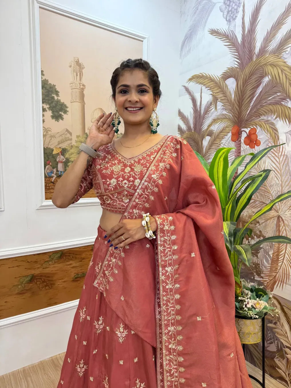 Woman in a traditional red saree with floral patterns indoors, with plants and artwork in the background.