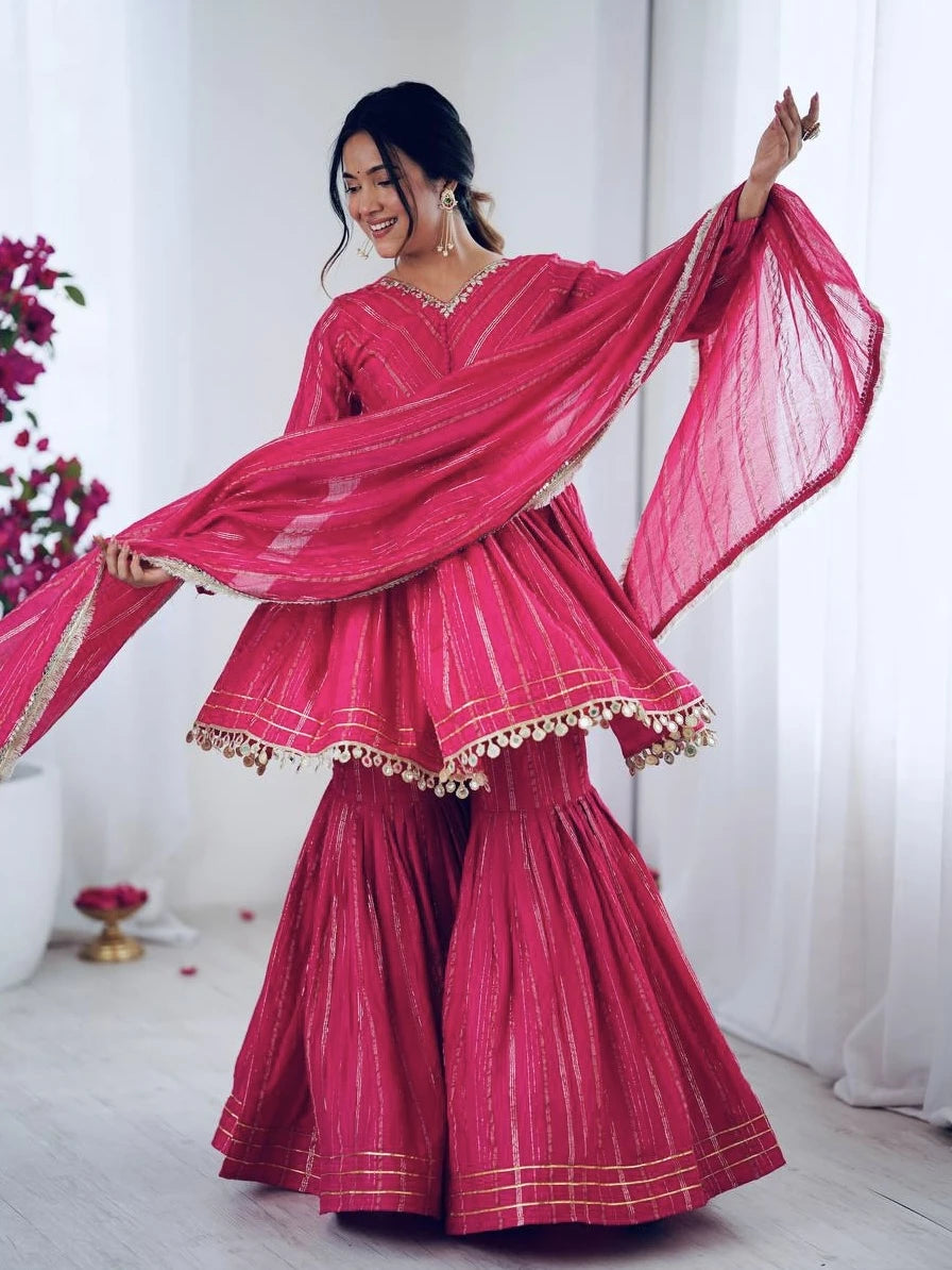 Woman in a pink traditional outfit with a dupatta, standing against a white curtain.