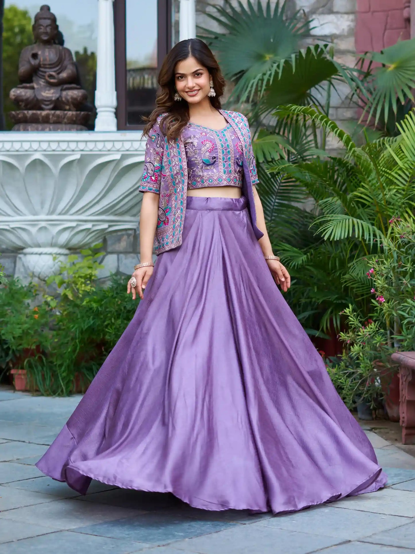 Woman in a purple traditional outfit standing outdoors with greenery and a statue in the background
