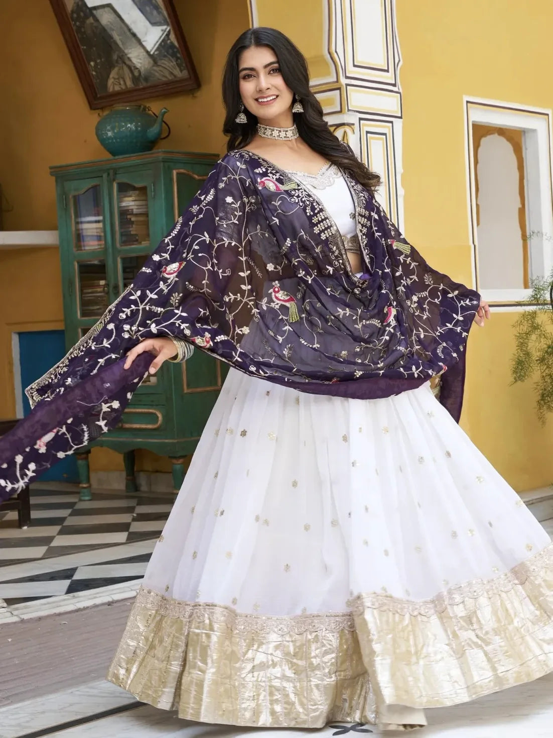 Woman in a white traditional outfit with a purple dupatta in a decorated room.