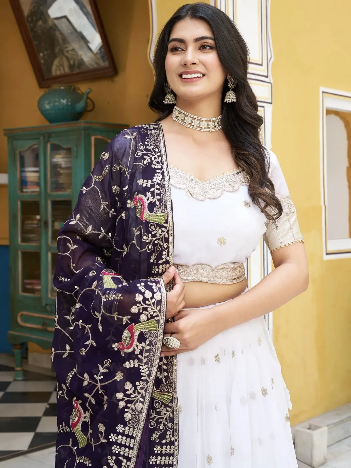 Woman in a white outfit with a purple embroidered dupatta against a yellow wall.