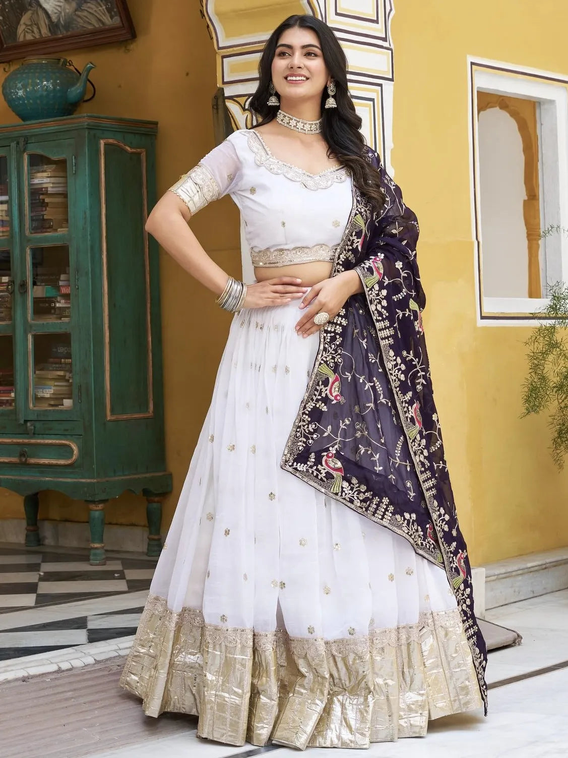 Woman in a white and gold traditional outfit with a dark purple dupatta in an indoor setting.