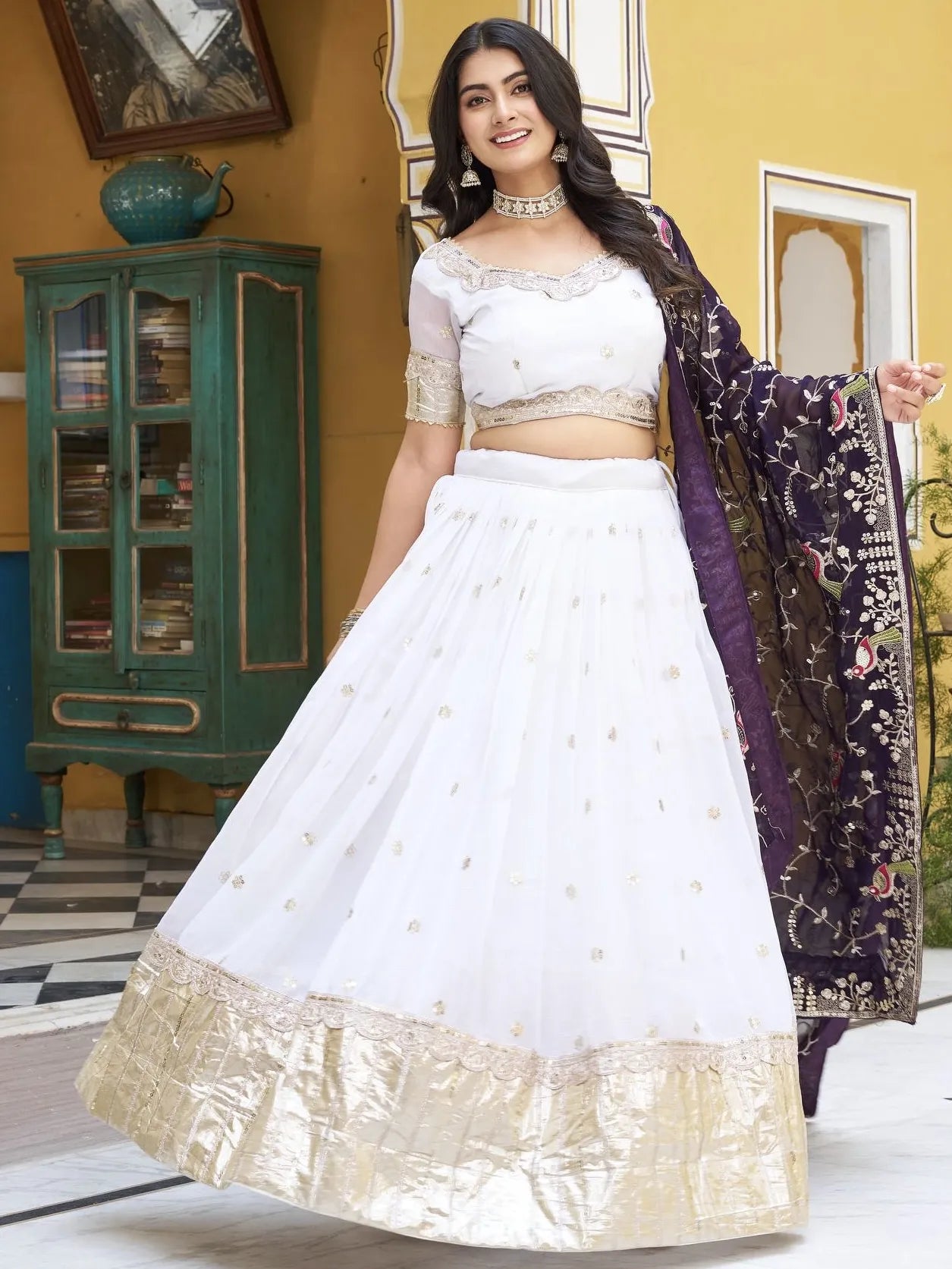 Woman in a white traditional outfit with gold details, holding a purple and gold shawl.