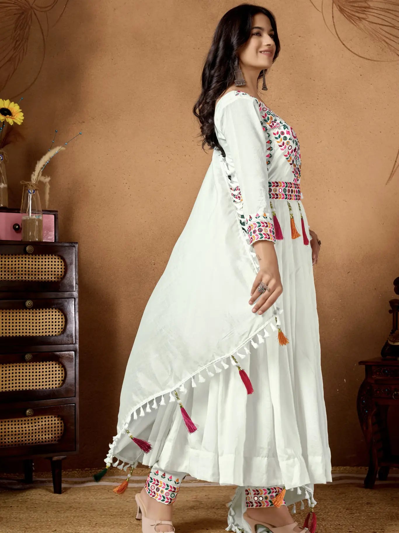 Woman wearing a white embroidered dress with colorful patterns in a warm-toned room.