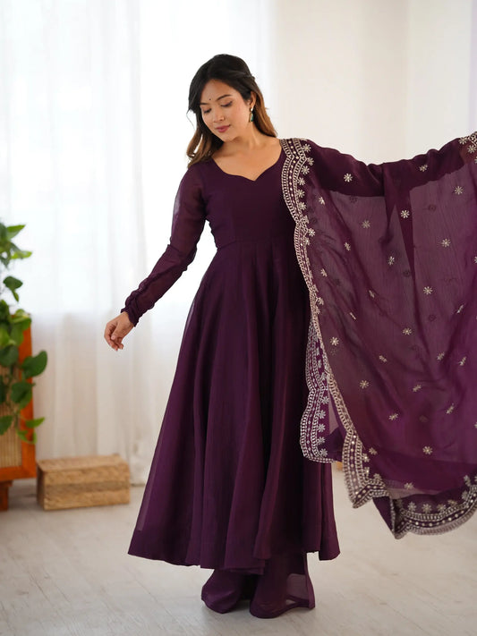 Woman wearing a long purple dress with a matching dupatta in a room with white curtains and a plant.