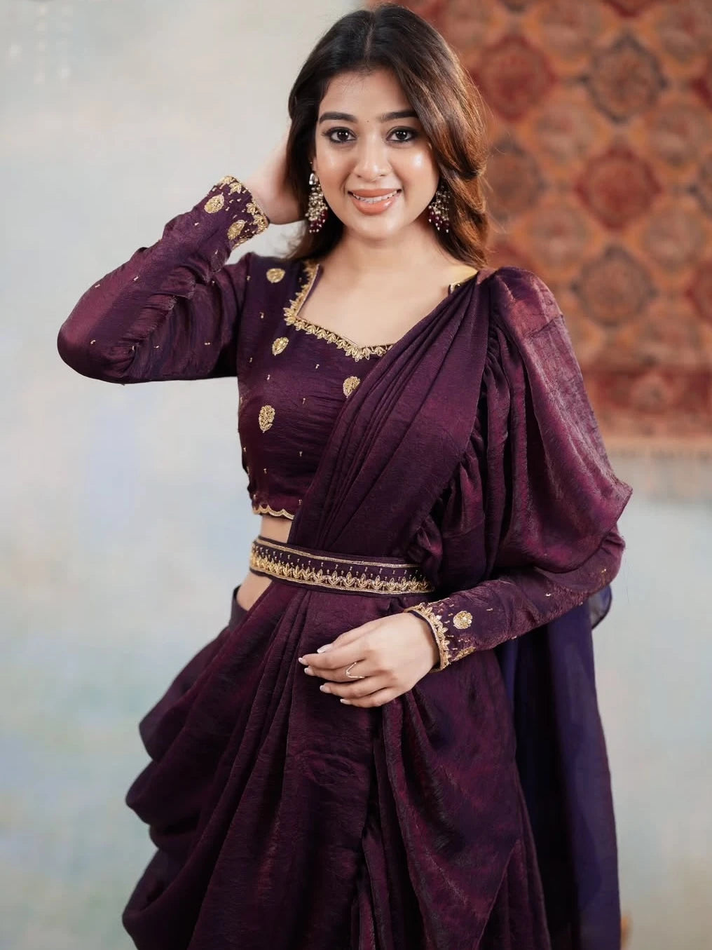 Woman wearing a dark purple traditional outfit with gold embroidery against a patterned wall.