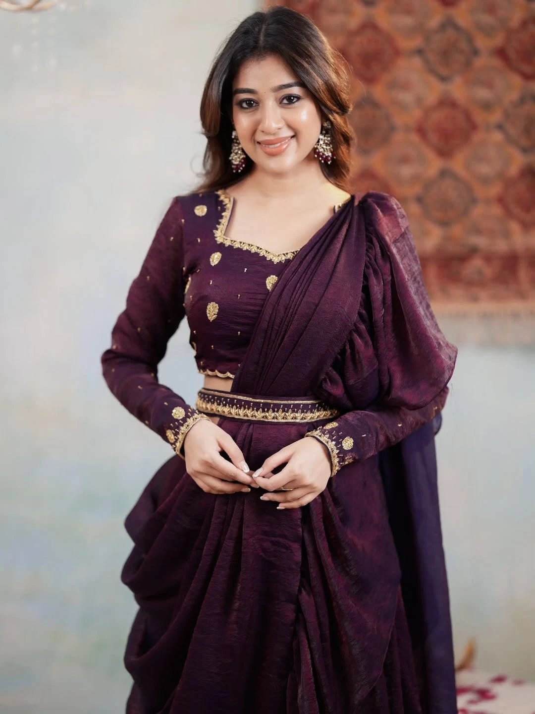 Woman wearing a dark purple saree with gold accents against a patterned wall.
