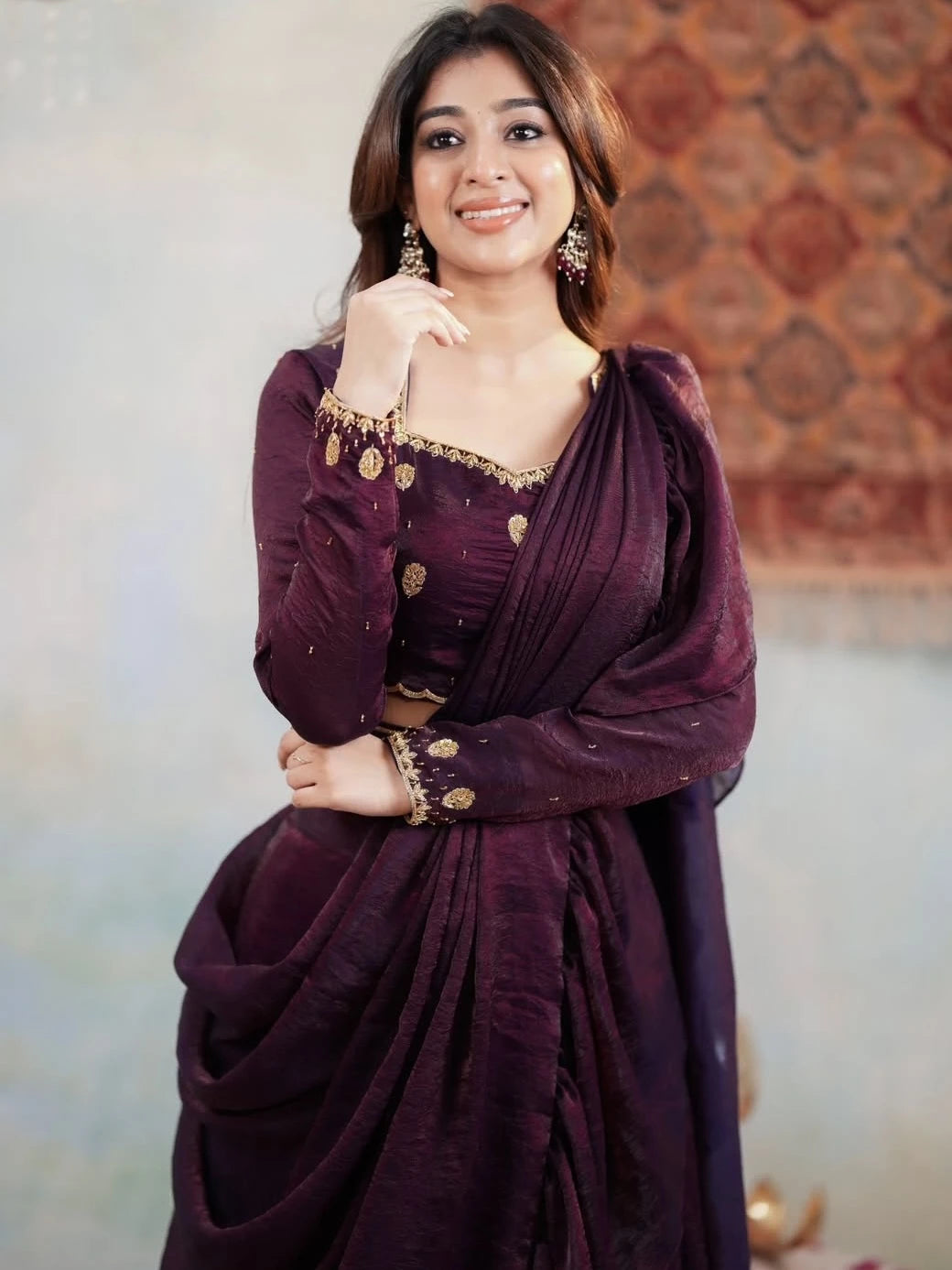 Woman wearing a purple saree with gold accents against a patterned wall.