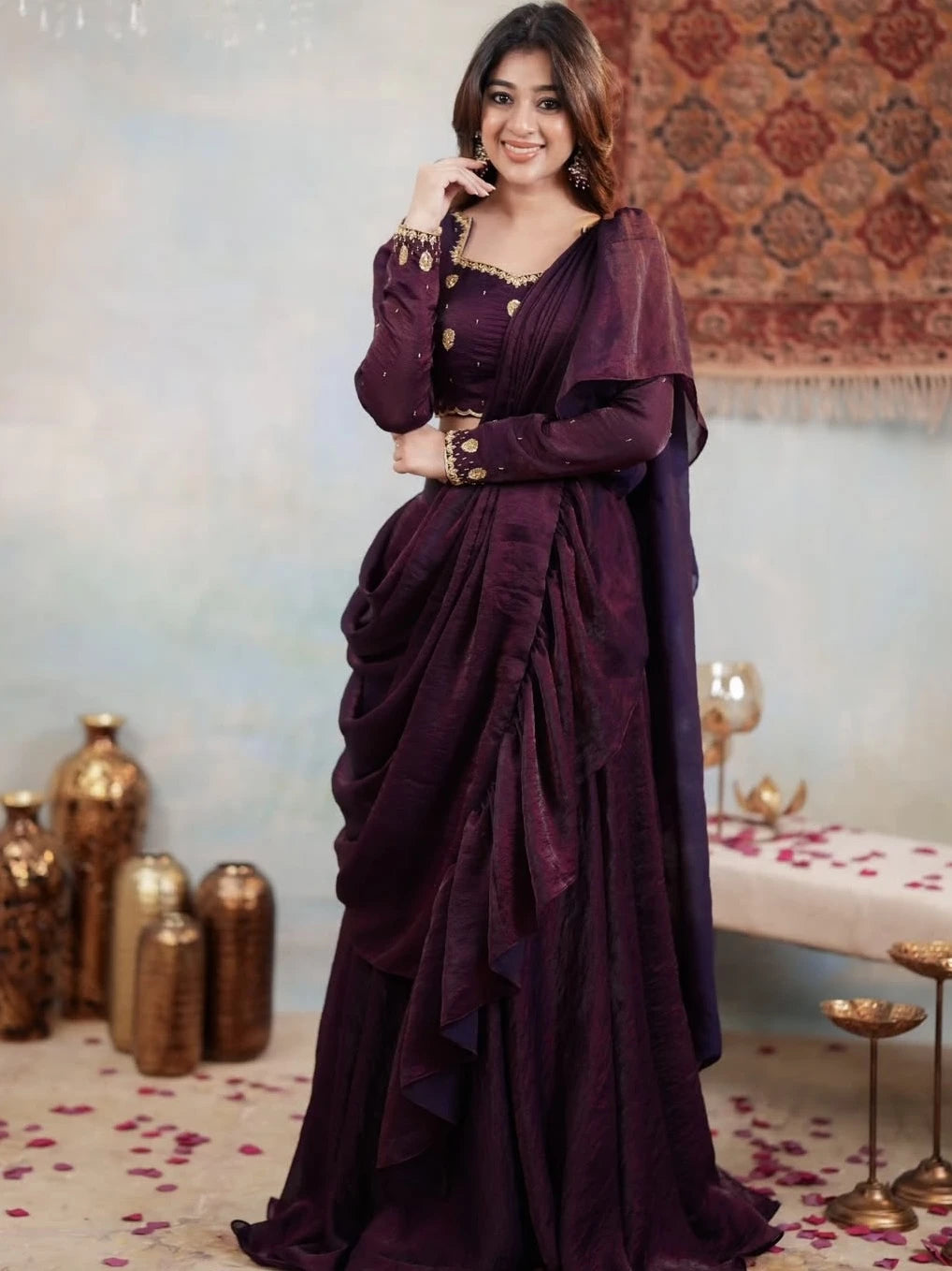 Woman in a purple saree with gold accents standing in a decorated room.