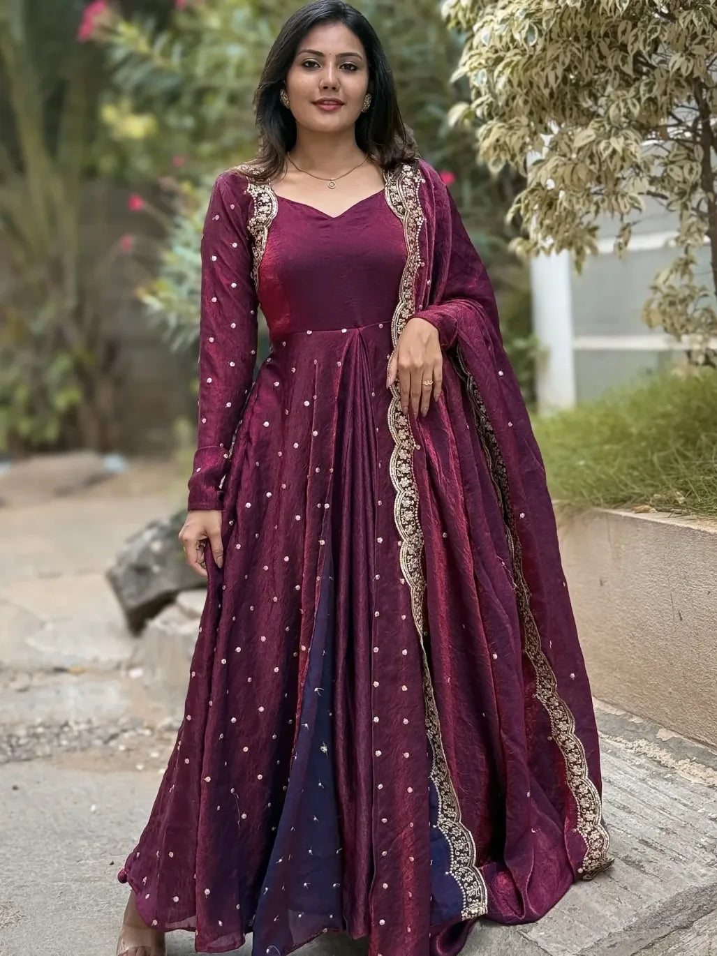 Woman wearing a maroon traditional outfit with gold embroidery outdoors.