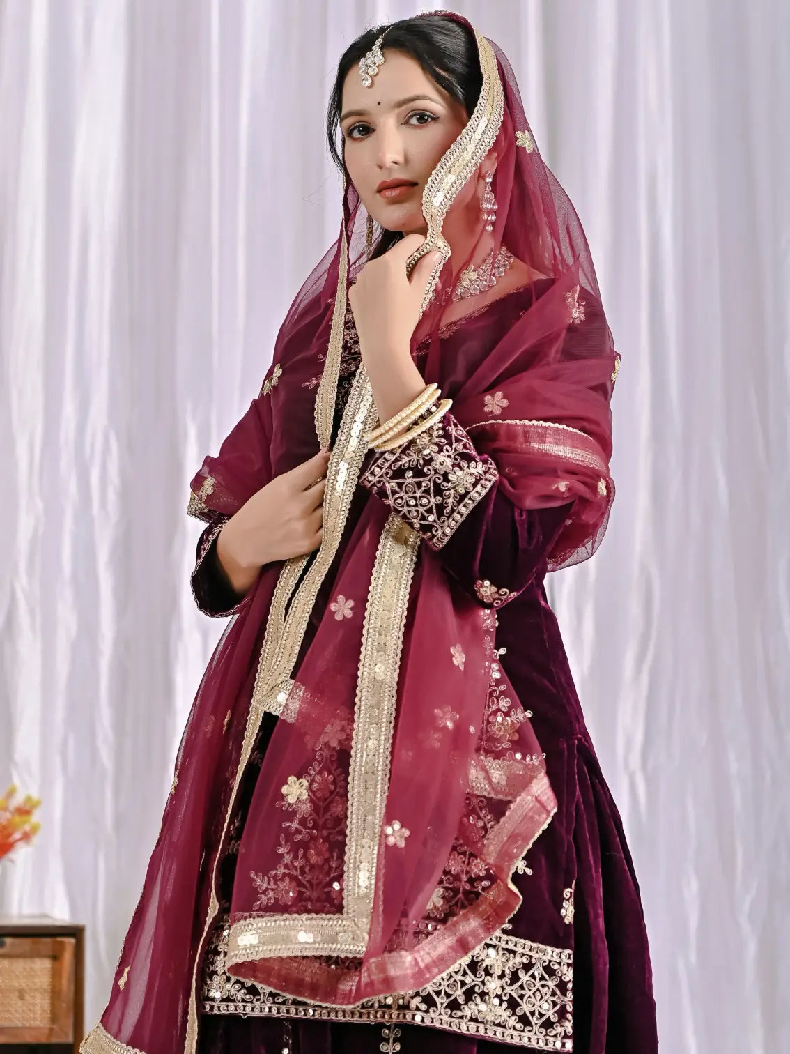Woman wearing a traditional wine and gold outfit with a dupatta against a white curtain background
