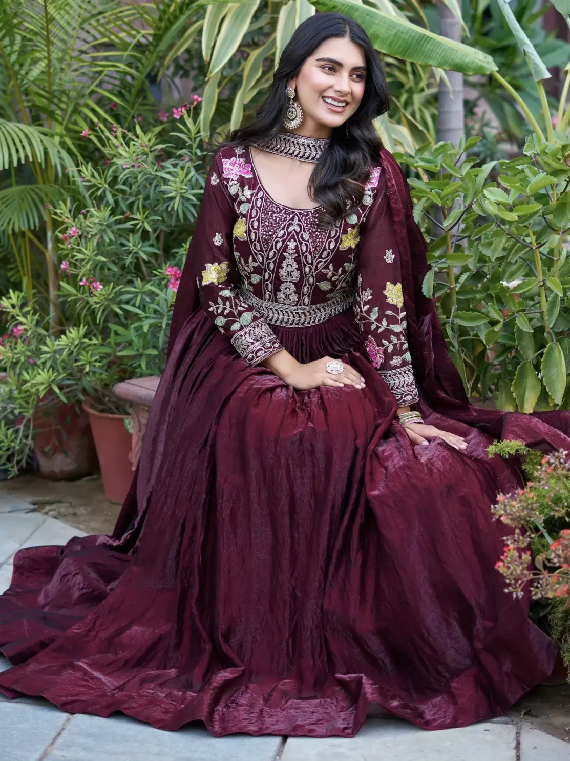Woman in a burgundy embroidered traditional outfit sitting outdoors with greenery.