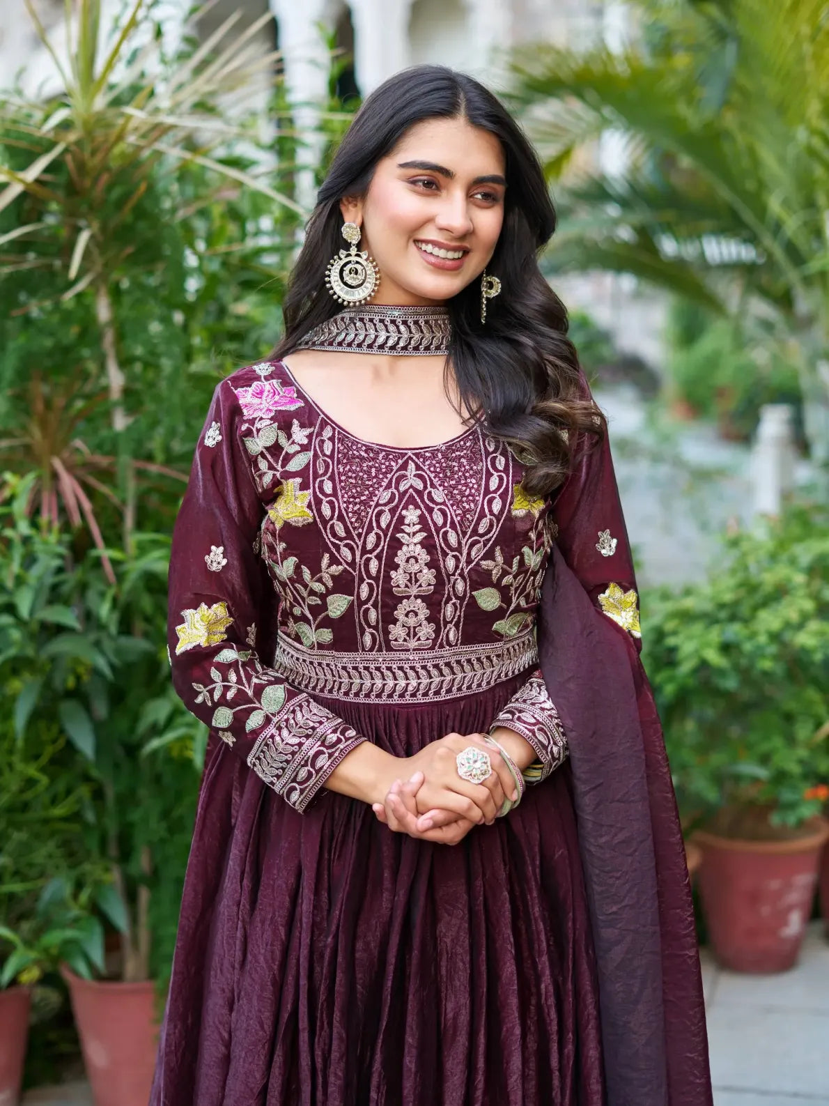 Woman in a maroon embroidered traditional outfit standing outdoors with greenery.